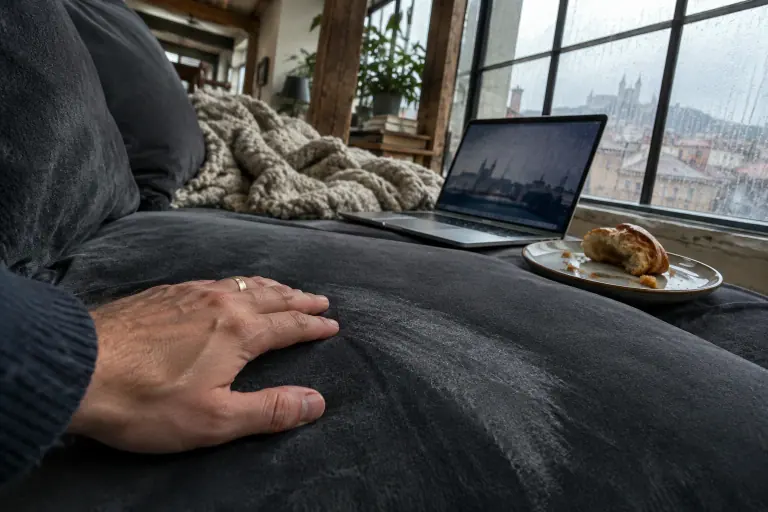 Gros plan sur une main qui caresse l’assise d’un canapé en velours gris anthracite, laissant une trace plus claire, dans un loft lyonnais un jour de pluie