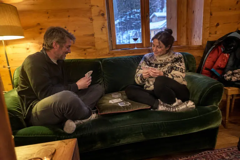 Un couple joue aux cartes assis sur un canapé en velours vert émeraude, dans un chalet en bois savoyard un soir de neige, sous une lampe allumée