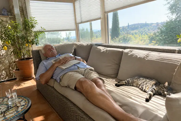 Un homme âgé fait la sieste sur un canapé en résine tressée grise à coussins sable, dans une véranda provençale plein sud, un chat allongé au soleil