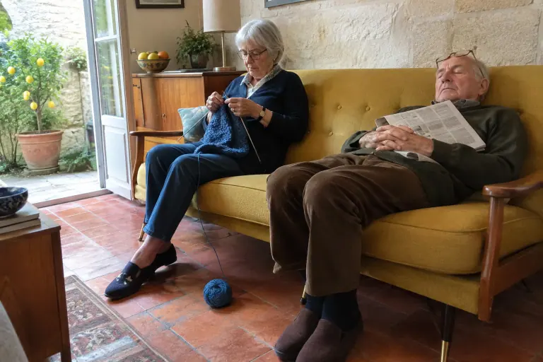 Un couple de septuagénaires sur un canapé vintage en teck et laine moutarde : elle tricote, lui somnole, dans une échoppe bordelaise au printemps