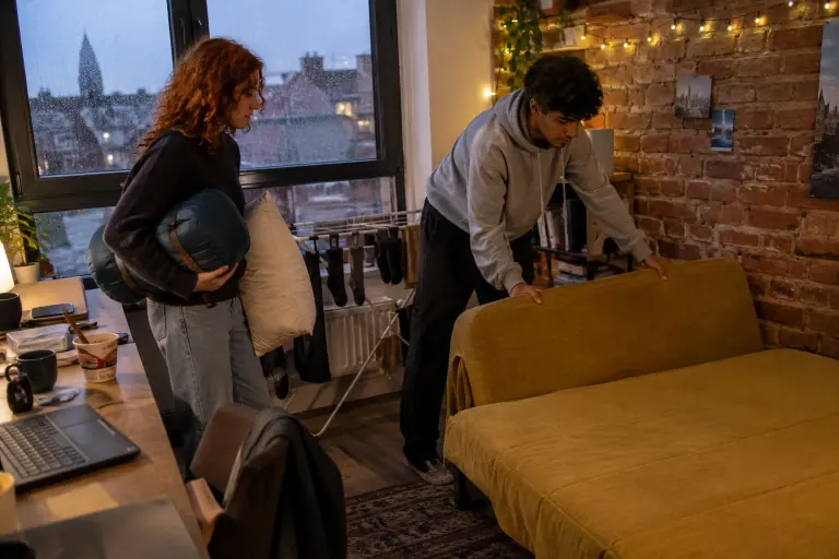 Dans un studio étudiant à Lille, un jeune homme abaisse le dossier d’un clic-clac jaune moutarde pour en faire un lit, son amie attend avec un sac de couchage