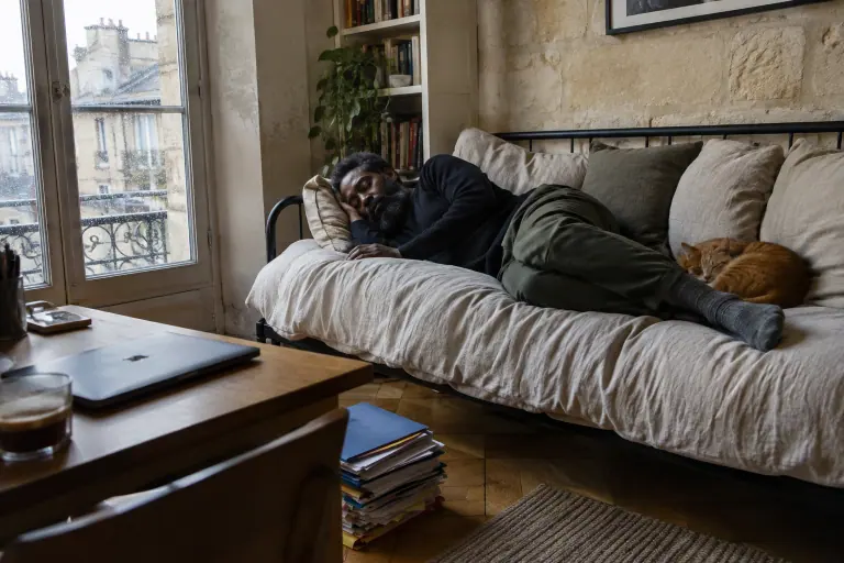Un homme fait la sieste sur un daybed en métal noir couvert d’un jeté en lin, un chat roux à ses pieds, dans un bureau bordelais un jour de pluie