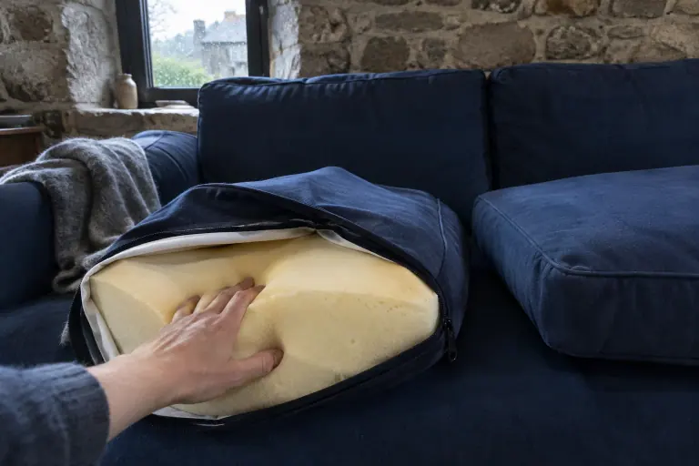 Une main s’enfonce dans la mousse jaune pâle d’un coussin d’assise à moitié déhoussé, posé sur un canapé bleu marine dans une longère bretonne un jour de pluie