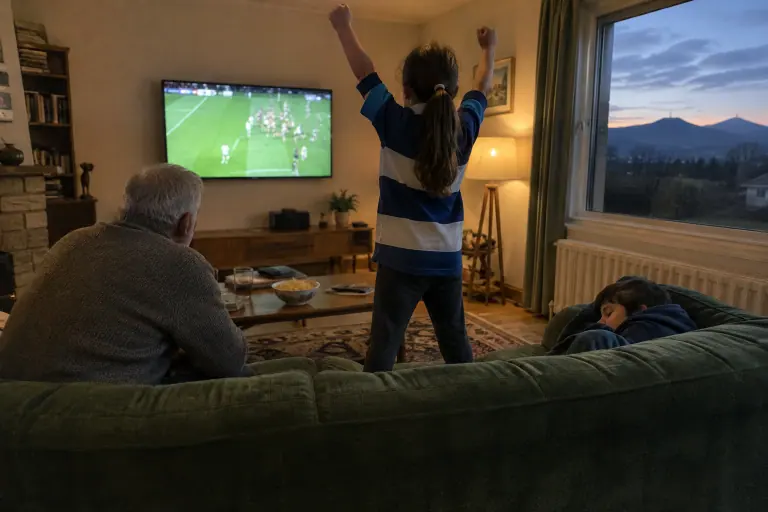 Vu de derrière un canapé vert foncé, un grand-père et ses petits-enfants regardent un match de rugby sur une télévision murale, lampe allumée