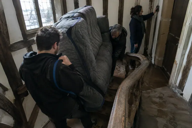 Deux hommes montent un canapé emballé de couvertures dans un escalier tournant en bois, pendant qu’une femme mesure l’encadrement de porte sur le palier