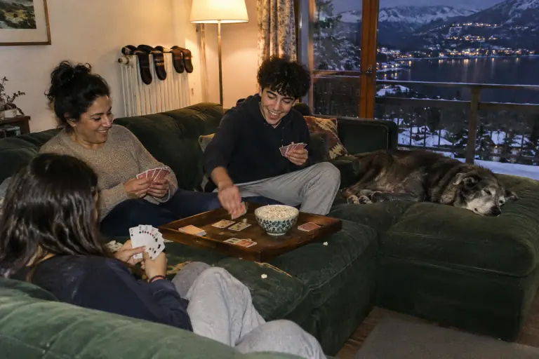 Une mère et ses deux adolescents jouent aux cartes sur un grand canapé d’angle vert sapin avec méridienne, un chien endormi, un soir d’hiver à Annecy