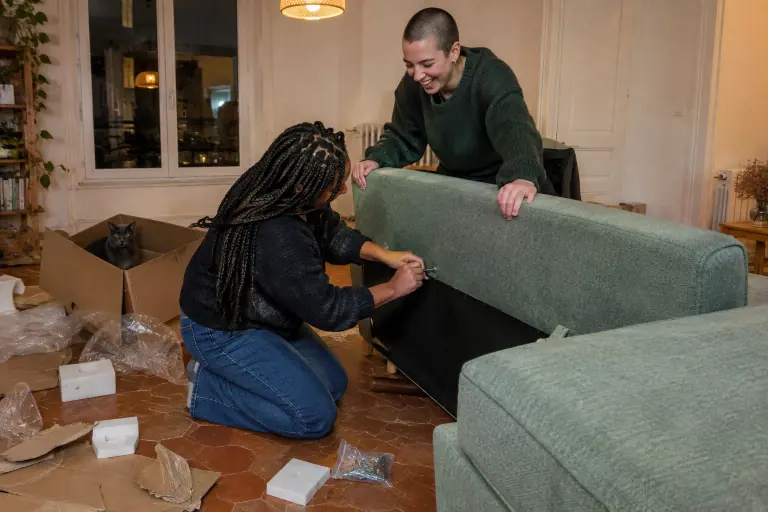 Deux jeunes femmes montent un canapé deux places vert sauge au milieu des cartons, l’une serre un boulon du dossier, un chat assis dans une boîte vide