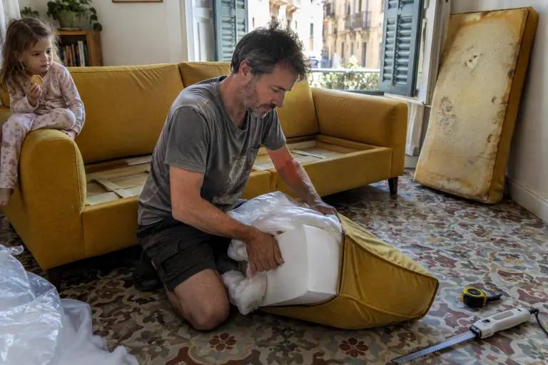 Un homme agenouillé glisse une mousse neuve entourée de ouate dans la housse d’un coussin de canapé moutarde, sa fille le regarde depuis l’accoudoir