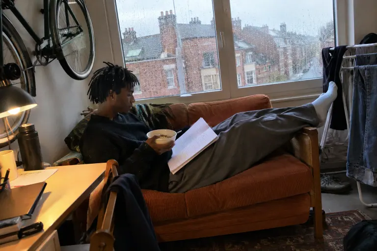 Dans un studio lillois, un étudiant mange ses céréales sur un mini canapé en velours côtelé terracotta, jambes sur l’accoudoir, sous une fenêtre pluvieuse