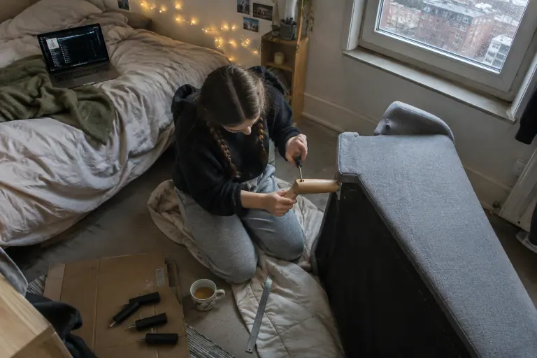 Une étudiante visse un pied conique en chêne sous un canapé gris deux places renversé sur une couette, dans un studio à Lille