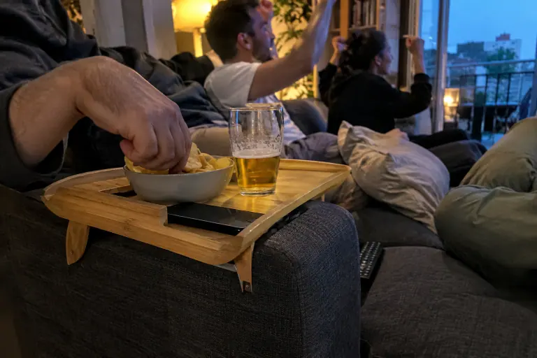 Un plateau en bambou posé sur l’accoudoir carré d’un canapé gris porte un verre, un téléphone et des chips, pendant un match entre colocataires