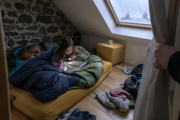 Deux fillettes en sac de couchage sur un pouf convertible moutarde déplié au sol, un second pouf encore fermé dans un coin de la chambre mansardée