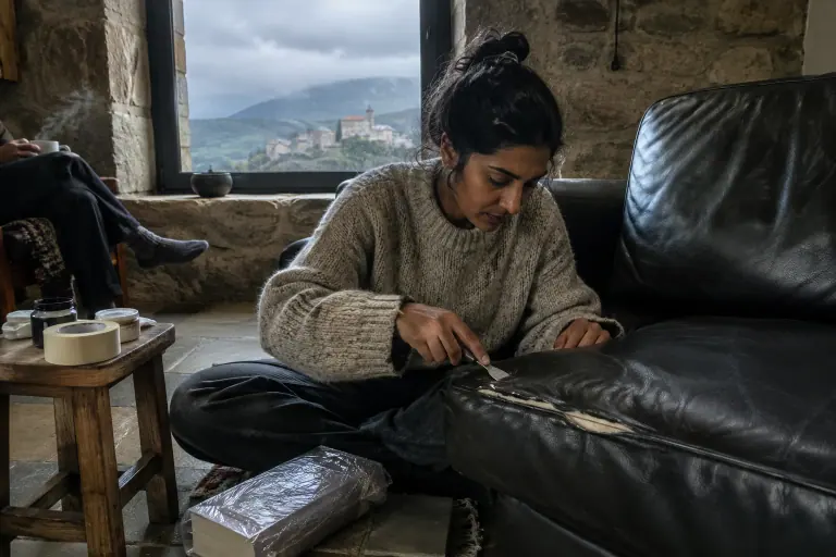 Une jeune femme assise par terre referme une déchirure sur l’assise d’un canapé en cuir noir avec une spatule, dans une maison de village en pierre