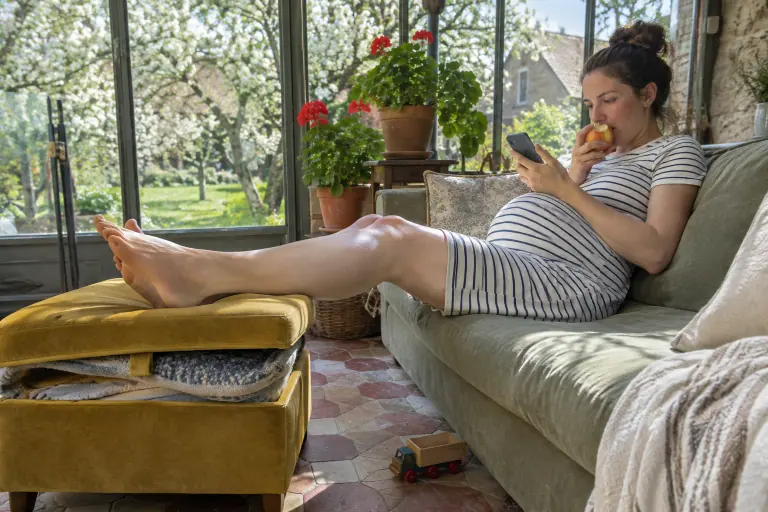 Une femme enceinte repose ses pieds sur un pouf coffre en velours moutarde devant un canapé vert sauge, dans une véranda normande au printemps