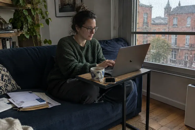 Une femme travaille sur son ordinateur posé sur une table en C glissée sous l’assise d’un canapé bleu marine, un jour de pluie à Grenoble