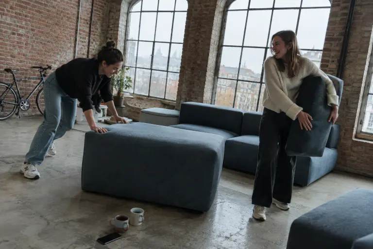 Deux amis déplacent les modules d’un canapé modulable bleu ardoise sur le sol en béton d’un loft lyonnais, un matin d’automne gris